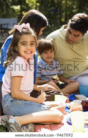Hispanic girl smiling at viewer with family picnicing in the park.