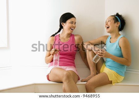 Two young women in fitness clothes holding water bottles sitting smiling and laughing and talking.