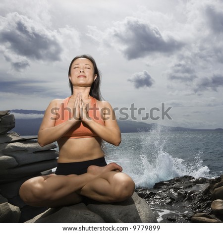 Asian woman sitting on rock by ocean in lotus pose with eyes closed in Maui, Hawaii