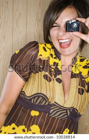 Caucasian mid adult brunette woman looking through miniature toy camera and making facial expression.