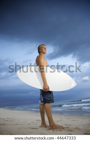 Young male surfer standing on beach with surfboard looking out at ocean.