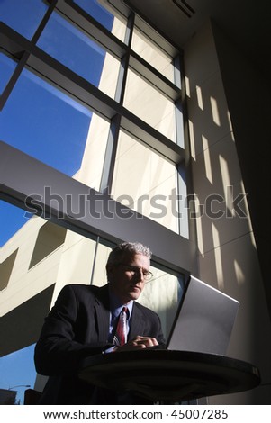 Low angle view of a businessman with a laptop computer in front of large windows. Vertical shot.