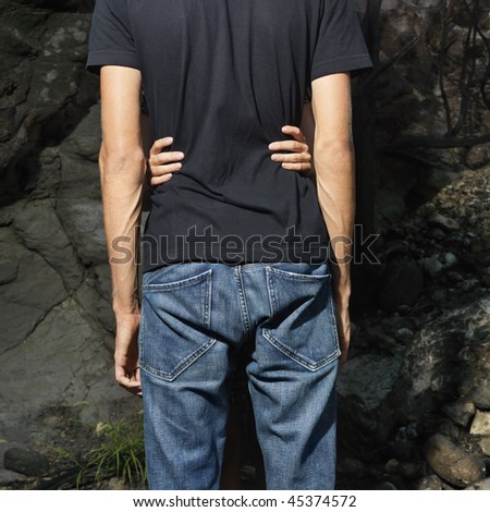 Rear view of a male in jeans and black t-shirt embraced by woman beside rocks. Square format.