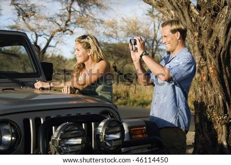 A woman leans on an SUV while a man uses a video camera to capture the scenery. Horizontal format.