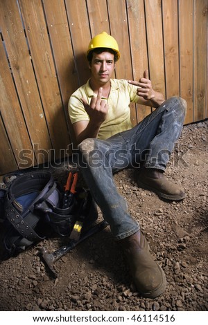 Male Caucasian construction worker in a yellow hard hat sits against the fence with his tools giving the finger. Vertical shot.