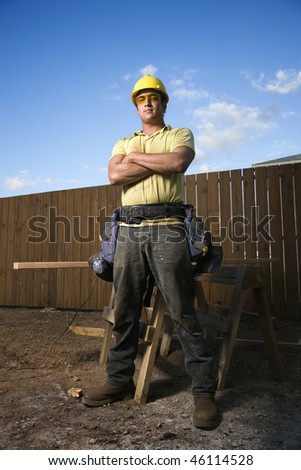 Male Caucasian construction worker stands confidently and looks into the camera. His arms are folded across his chest and a workbench can be seen behind him. Vertical shot.