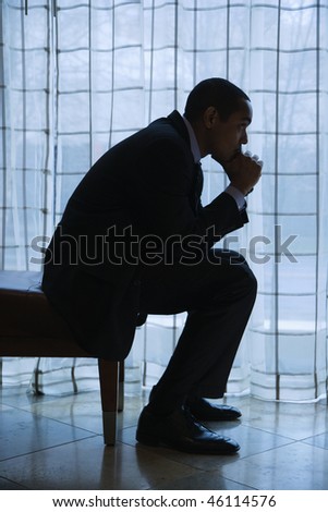 Profile of African-American mid-adult businessman sitting and thinking with hand on chin next to a curtained window. Vertical format.