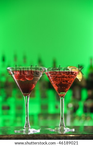 Two cocktails, one with a lemon wedge, sit on a bar counter against a green background. Veritcal shot.