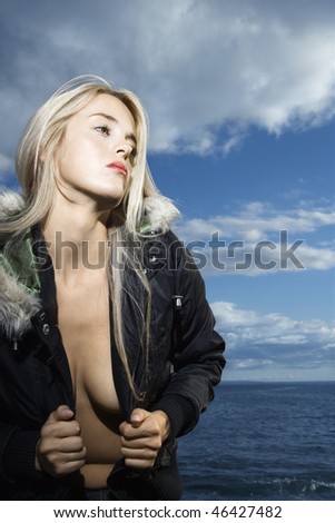 Attractive young woman opens the front of her parka exposing bare skin with the ocean in the background.