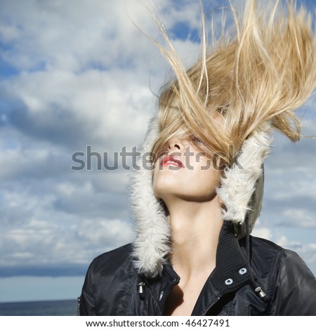 Attractive young woman is wearing a parka at the beach and flipping her long blond hair back. Square format.