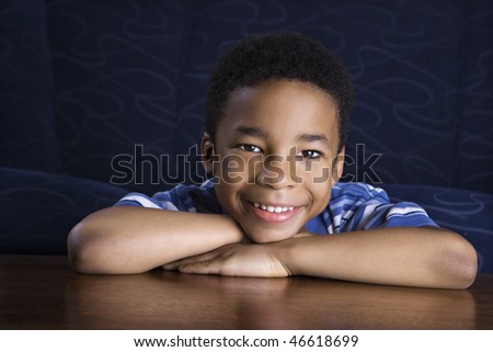Young African American boy sits at a coffee table with his chin propped on his hands as he smiles towards the camera. Horizontal shot.