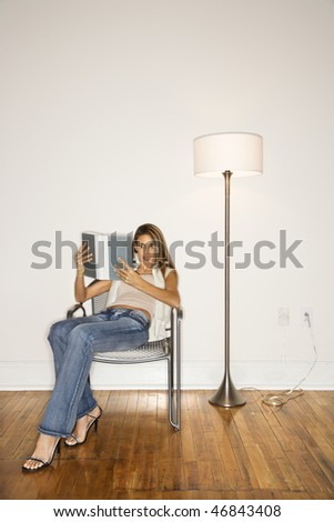 Attractive young woman sitting back in a silver chair and reading a book next to a floor lamp. Vertical shot.
