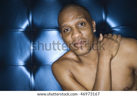 Handsome young man sitting with his hand on his shoulder against blue padded background, shirtless and smiling. Horizontal shot.