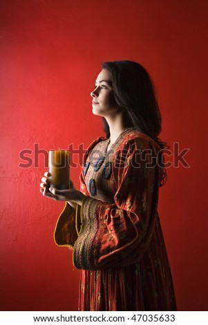 Attractive young adult woman in cultural dress, standing against red wall and holding a candle. Vertical shot. Isolated on red.