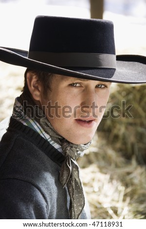 Attractive young man wearing a black cowboy hat with bales of hay in the background. Vertical shot.