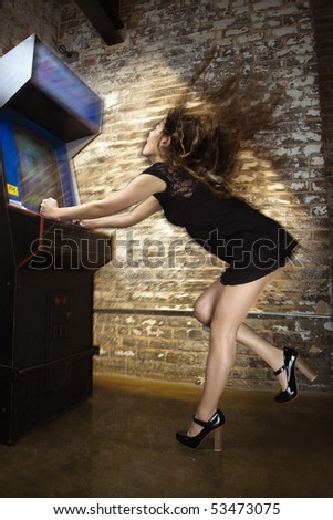 Attractive young woman standing wearing short dress playing video arcade game with hair flying out behind her.