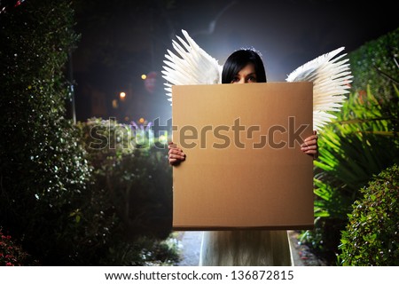 Angel woman with white wings holding blank cardboard message board poster in night garden. Copyspace.