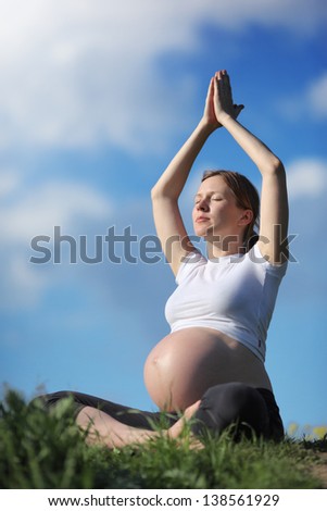 Pregnant woman practicing yoga in lotus pose on green grass  under blue sky