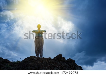 Woman standing on top of mountain doing yoga meditation. Stormy sky background, sun rays shining on her.
