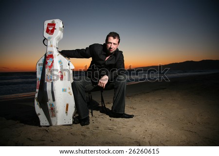 Portrait of a musician with travel cello or guitar case, sitting on beach at night. Shallow DOF.