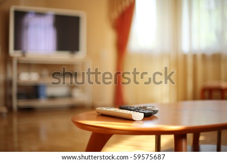 Romote controls on wooden table, tv in the background. Shallow DOF, focus on white remote.