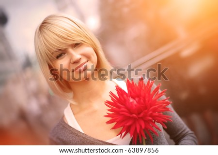 Portrait of beautiful young Caucasian woman with red flower smiling over city street background. Closeup, shallow DOF.
