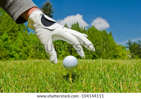 A hand in the golf glove placing golf ball on the tee with  nice golf course and  blue sky as a background