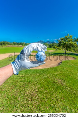 Hand wearing golf glove holding golf ball over beautiful golf course with blue sky. Vertical.