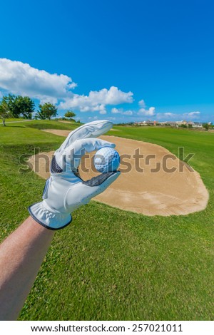 Hand wearing golf glove holding golf ball over beautiful golf course with blue sky.