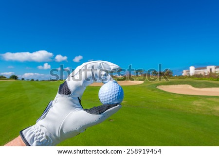 Hand wearing golf glove, holding golf ball over beautiful golf course with sand bunkers at the back.
