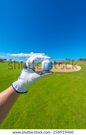 Hand wearing golf glove, holding golf ball over beautiful golf course with sand bunkers at the back. Vertical.