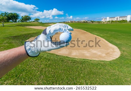 Hand wearing golf glove holding golf ball over beautiful course with blue sky.