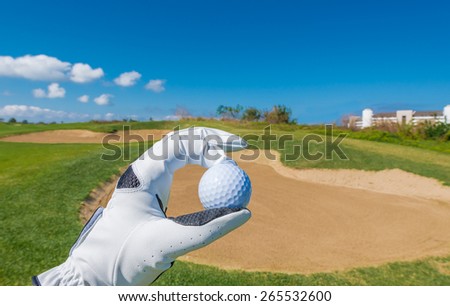 Hand wearing golf glove holding golf ball over beautiful course with blue sky.