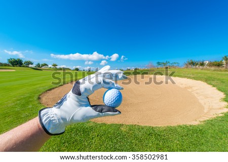 Hand wearing golf glove, holding golf ball over beautiful golf course with sand bunkers at the back.