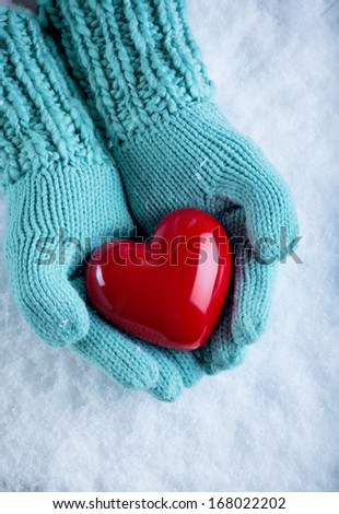Woman hands in light teal knitted mittens are holding a beautiful glossy red heart in a snow background. Love and St. Valentine concept.