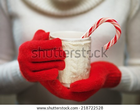 Woman holds a winter cup close up. Woman hands in woolen red gloves holding a cozy mug with hot cocoa, tea or coffee and a candy cane. Winter and Christmas time concept.