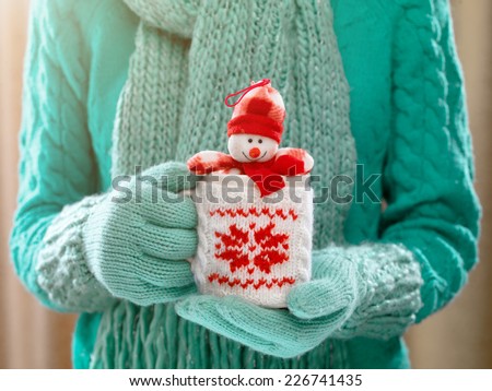 Woman holding winter cup with nice Christmas toy close up on light background. Woman hands in woolen teal gloves holding a cozy mug with happy snowman toy. Winter and Christmas time concept.
