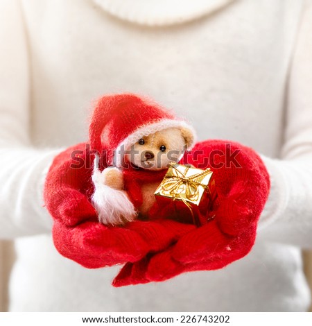 Female hands holding a cute teddy bear. Woman hands in red mittens showing a teddy bear gift dresses in knitted hat and scarf. Cute Christmas present. Winter holidays concept.