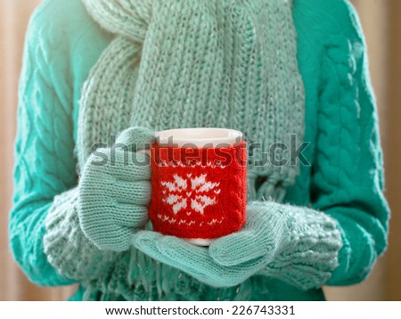 Woman holding winter cup close up on light background. Woman hands in teal gloves holding a cozy mug with hot cocoa, tea or coffee and a candy cane. Winter and Christmas time concept.