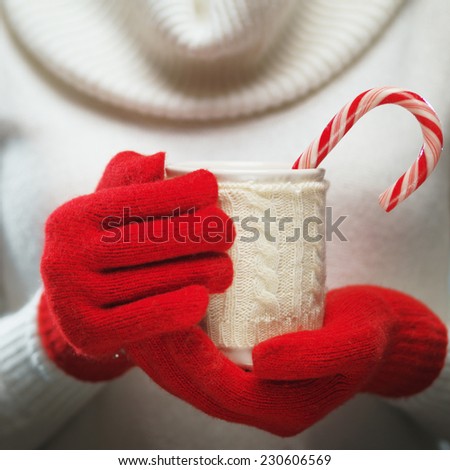 Woman holds a winter cup close up. Woman hands in woolen red gloves holding a cozy mug with hot cocoa, tea or coffee and a candy cane. Winter and Christmas time concept.