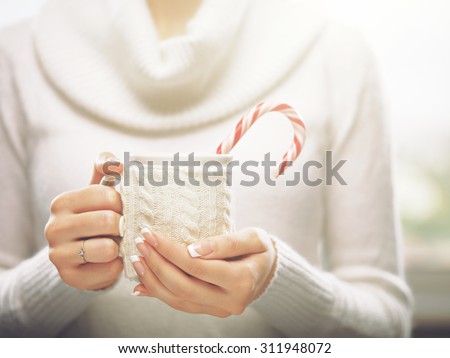 Woman holds a winter cup close up. Woman hands with elegant french manicure nails design holding a cozy knitted mug with cocoa, tea or coffee and a candy cane. Winter and Christmas time concept.