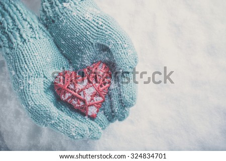 Woman hands in light teal knitted mittens are holding a beautiful glossy red heart in a snow winter background. Love and St. Valentine concept.