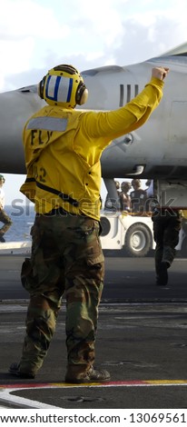 A Navy sailor directs an F-18 fighter aircraft around the flight deck on an aircraft carrier