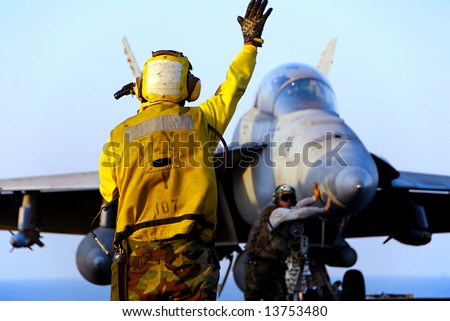A Navy Sailor directs an F-18 Hornet fighter aircraft around the flight deck of an aircraft carrier