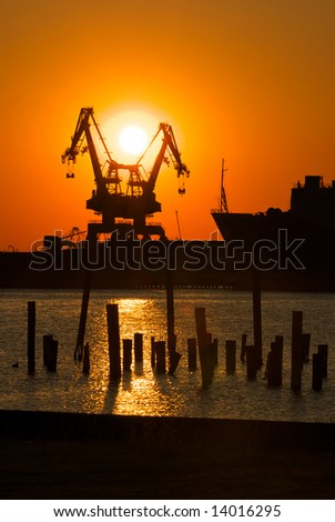 Two cranes sit dramatically against a colorful sunset in a large shipyard with old dock pilings in the foreground
