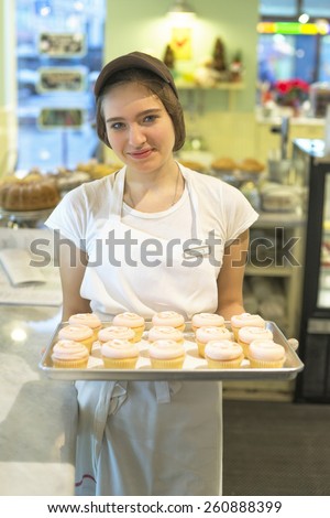 Smiling shy girl in the cap with cupcakes at the tray in the bakery