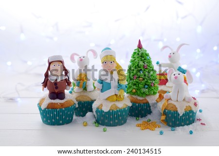 Festive cupcakes with sugar figures on a bright lights background. A kid in a fur hat Snow Maiden and rabbits decorating the Christmas tree.
