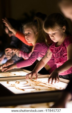 Girl paint with sand on table by her fingers in classroom with friends