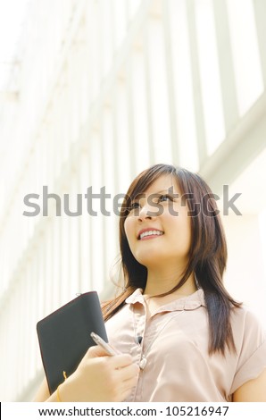 A young Asian woman looking far away to bright light in front of a modern office building, with diary on hand.