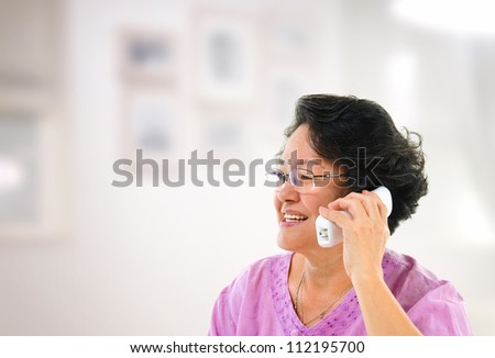 Happy mature Asian woman talking on phone, inside house